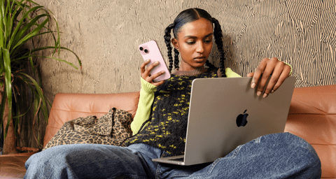 Osoba, která sedí na pohovce, v pravé ruce drží iPhone a na klíně má otevřený MacBook