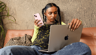 Osoba, která sedí na pohovce, v pravé ruce drží iPhone a na klíně má otevřený MacBook