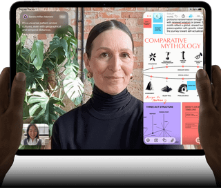 iPad Pro, pohled zepředu, orientace na šířku, vesmírně černá barva, zaoblené rohy, černý rámeček displeje, ruce drží levou a pravou stranu, na displeji se zobrazuje FaceTime konverzace překládaná v reálném čase a vzdělávací aplikace s prezentací na téma komparativní mytologie, textem, diagramy a ručně psanými poznámkami