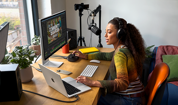A woman works at home at her desk on a MacBook Pro connected to two external monitors, wearing headphones and speaking into a microphone.